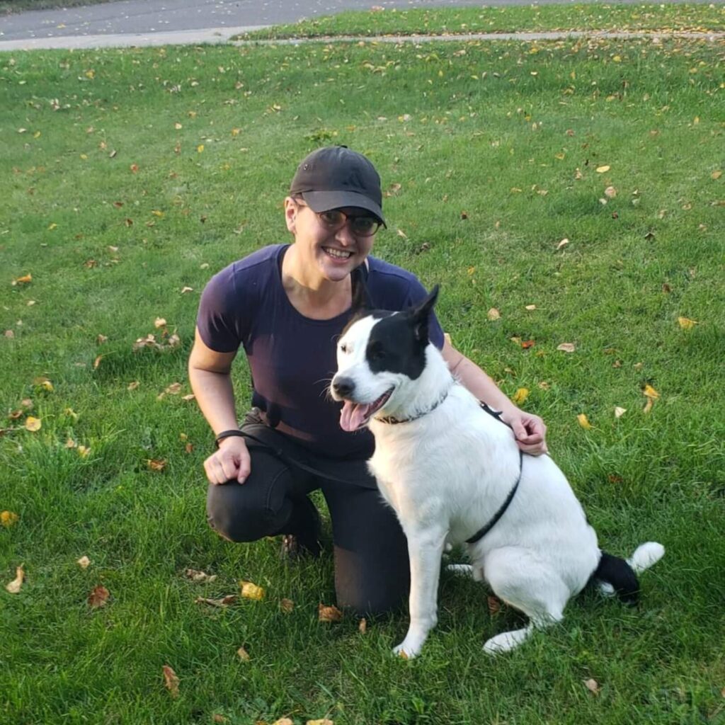 Traci Scott, female in black baseball cap and casual attire, kneeling on ground with large white and black dog.